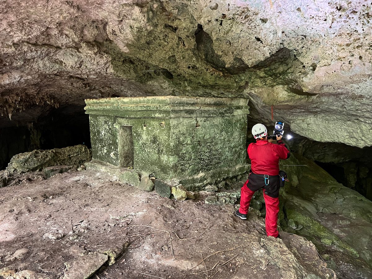 A researcher in a red jumpsuit scans a small Maya shrine inside a cave.