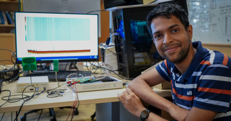 Graduate student Raghav Subbaraman smiles beside a new electronic device for more accessible, efficient wireless communications.